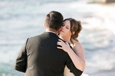 Bride and Groom holding each other close. Bride facing camera, Groom facing away towards water