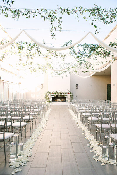A bright, elegant ceremony setup at SoHo63, a Chandler wedding and event venue, featuring rows of silver chiavari chairs, a petal-lined aisle, and white draping with greenery suspended overhead. A floral-decorated fireplace serves as the romantic focal point.