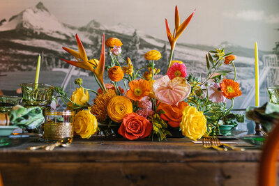 Vibrant arrangement of anthurium, heliconia, roses and ranunculus in orange, yellow and pale pink on a dark wood table at Campfire Hotel in Bend Oregon