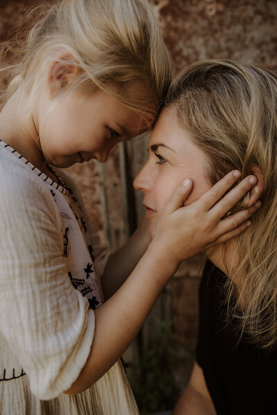 Closeup of mother and child, forehead to forehead. The child is standing up, the mother is crouching down and the child holds the mother's face in their hands.  