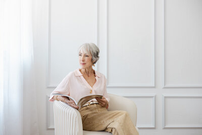 Woman wearing light pink blouse with tan pants holding a magazine and looking out the window against a white wainscotting wall in a Bay Area  brand photo studio.