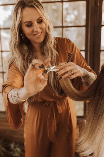 At the professional hair extensions and color salon the artist is cutting the hair of one of her clients. Behind her is a wood-framed window and she is dressed in an orange outfit.