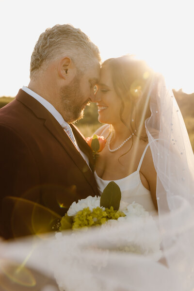 Bride and groom stand forehead to forehead during golden hour, wrapped in soft light and veil glow.