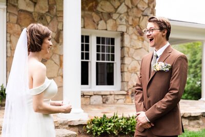 A groom bursts into joyful laughter during a first look with his bride, who smiles warmly while holding a small note in her hand. She wears an off-the-shoulder white gown with a veil, while he wears a brown suit with a floral boutonnière, standing together in front of a stone building.