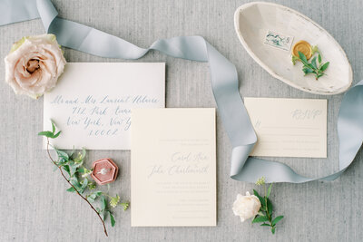 Calligraphy wedding invitations styled with blue ribbon, floral roses, and blush ring box