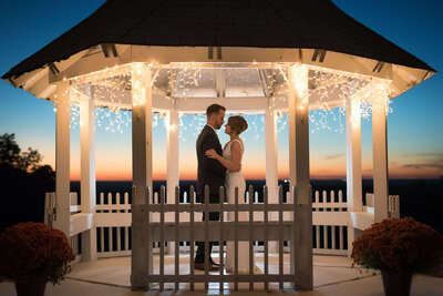 Bride and groom dance together under string lights inside Gazebo at sunset