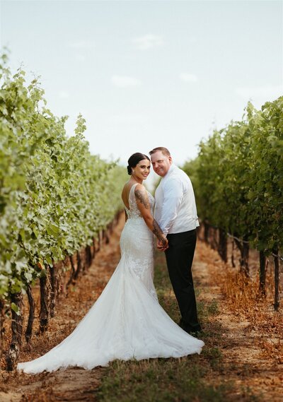 a couple facing away and looking into the camera. the bride is wearing a white long wedding dress and the groom is wearing a white shirt and black pants. They are standing in a lush green vineyard  at Covert Farms in Oliver
