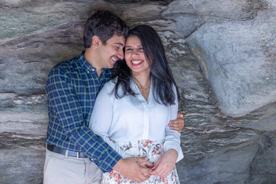 Bride to be in pale cream blouse and floral skirt being hugged by groom to be in blue and green striped shirt under rock overhang taken by top raleigh wedding photographer