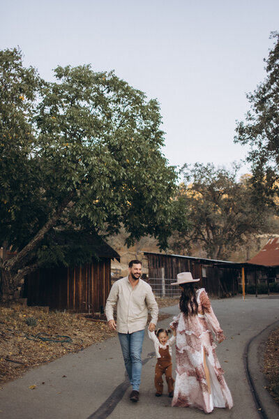 A family of three, including a young child, walking hand in hand along a rustic pathway surrounded by trees, with a barn in the background in a family photoshoot.