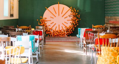 A wedding ceremony setup with a round pink and orange backdrop covered in marigolds and mismatched colorful seating