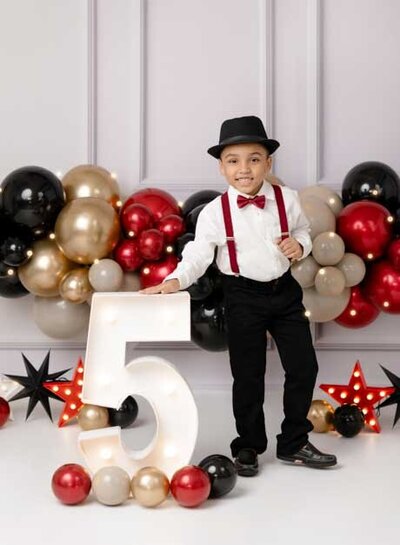 5th Birthday photo of boy in suspenders with red, black and gold balloons. Studio photo taken by Jenni Nikki at Woodstock Photo Studio.