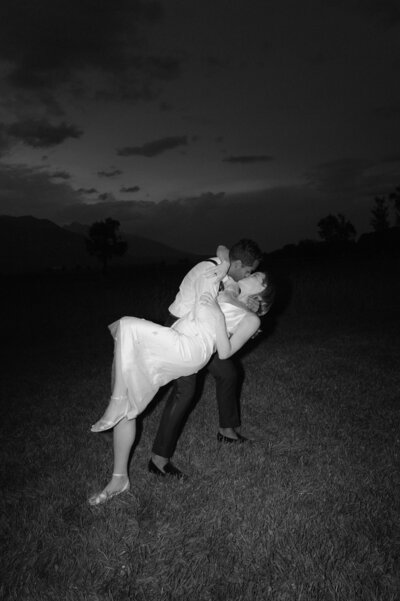 A black and white photo of a couple kissing in a dip pose outdoors at night, with a grassy field and trees in the background.