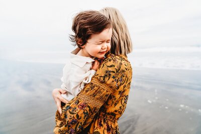 A toddler boy faces over his mother's shoulder as he scrunches his eyes shut adorably. They are standing on a San Diego beach during a family photoshoot.