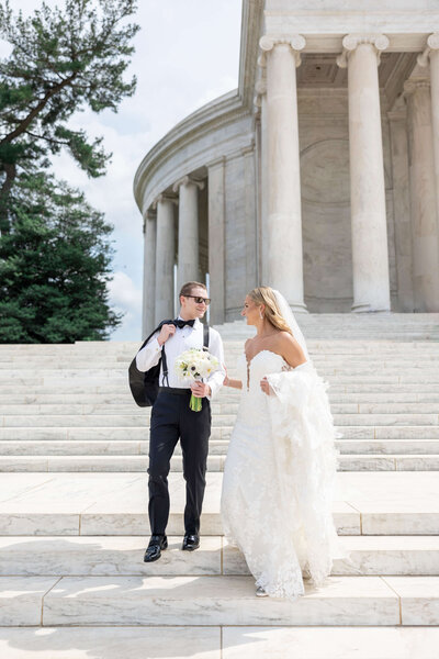 A bride and groom smile at each other on the steps of a grand building. The bride holds a white bouquet and wears a long, textured gown. The groom is dressed in formal attire with suspenders and carries a jacket over his shoulder.