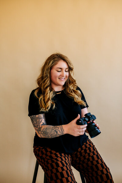 Woman sitting in a studio with solid background starting down at her camera smiling 