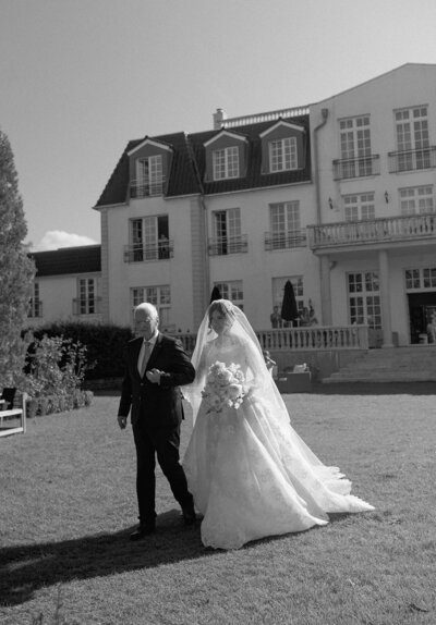 Black and white image of a bride walking down the aisle with her father-in-law at a wedding ceremony, wearing an Elie Saab dress.