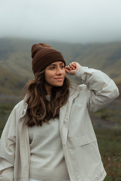 Smiling woman Orlando photographer sitting on a beige lounge chair holding a professional camera, dressed in a black blazer, beige pants, and white sneakers, with natural light streaming in at a studio in Orlando, Florida.