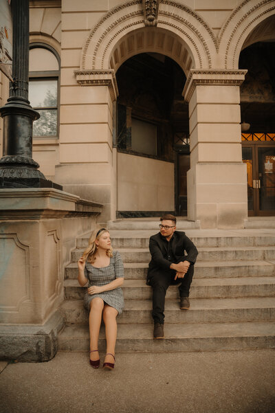 engaged couple sit on the steps of the museum in downtown bloomington illinois during their engagmeent session. the girl is wearing a vintage inspired dress while the man is in all black