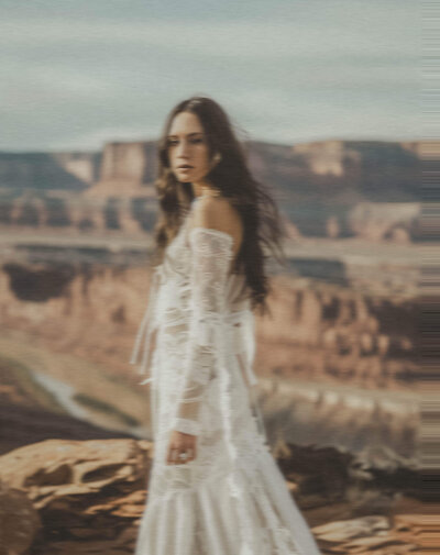 A woman in a long white dress stands outdoors on a rocky ledge with a canyon landscape in the background, embodying the elegance and creativity that inspires Sweet Pea Ink Creative's approach to brand design.