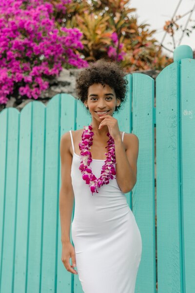 girl in flower lei by turquoise fence with pink flowers behind her