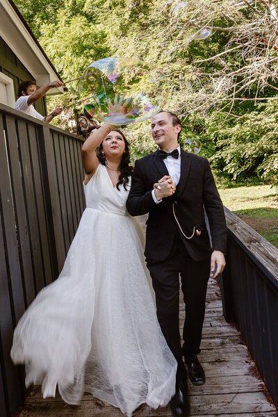 Two marriers on their wedding day laugh and hold hands, popping a bubble.