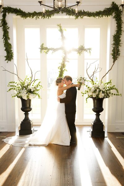 Gorgeous bride and groom pose in front of window with cross made of greenery