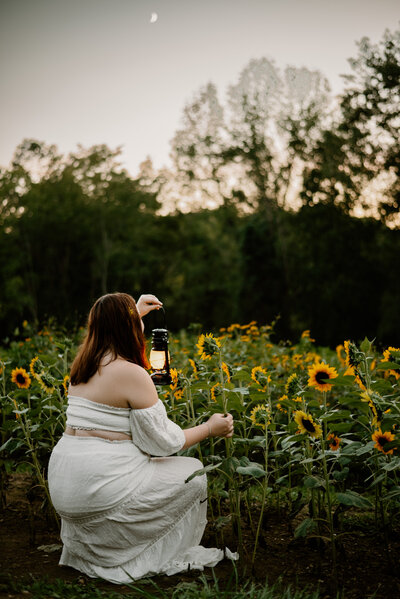 Teen girl in white dress crouched down with lantern in sunflower field with moon shining in Westminster Maryland.