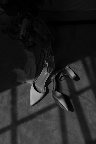Black and white photo of wedding shoes with a pointed toe, sprawled under the playful shadows and spills of light