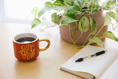 Red mug of black coffee with slogan Go Get 'Em on pale table with a potted plant, open blank notebook, and black pen. Photo by Kyle Glenn via Unsplash.