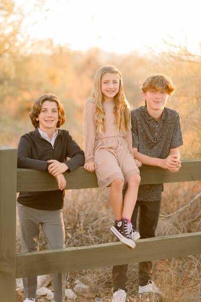 3 kids from a family smiling at the camera posed on a fence post