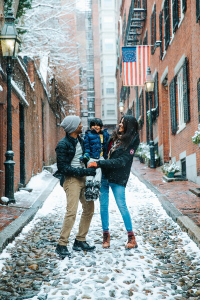 Family of 3 standing on a cobblestone street in Beacon Hill, Boston. Mom and dad are laughing holding their 6 year old in between them, child is looking at the camera. There is a brick building behind them with black shutters and an old gas streetlamp on their left.