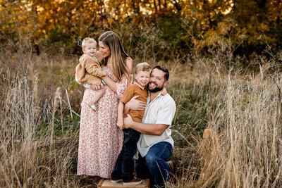 Family of four standing in the fall foliage embracing each other in hugs and smiling.