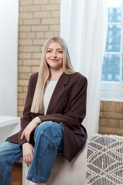 Steph, a young white woman with long blonde hair sitting on a stool. She is smiling. She is wearing a brown jacket and jeans