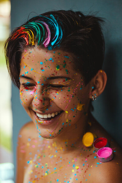 This image shows a young person, smiling widely with eyes close. They have streaks of different color paint in their hair, and their face and shoulders are covered in splatters of the same rainbow paint.