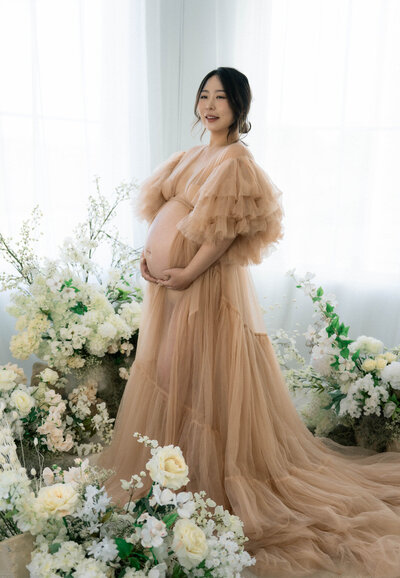 Floral maternity session in studio with mom in flowing open belly gown surrounded by florals
