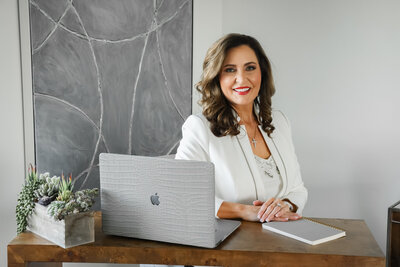 Woman in a white blazer sitting at a desk with a laptop and notebook, smiling at the camera. A potted plant and a grey abstract painting are in the background.