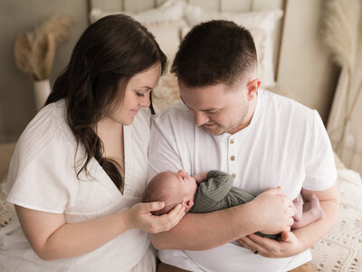mom and dad dressed in white sitting on bed holding newborn baby boy in green onesie for studio session