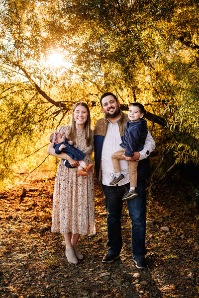 A family of four pose for a photo in front of a tree, the sun streaming through the yellow leaves