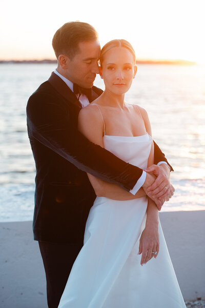 bride and groom embrace by water at sunset