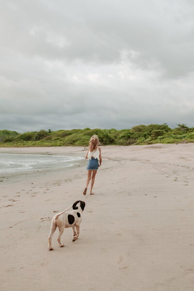 a girl i photographed on a beach in Costa Rica having her puppy follow her on a empty beach with the jungle and beach in the backdrop