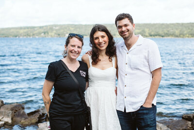 A smiling bride in a white lace dress and groom in a white button-up shirt pose with their wedding planner, wearing a "Home Town" shirt, by a scenic lakeside with blue water and green hills in the background.