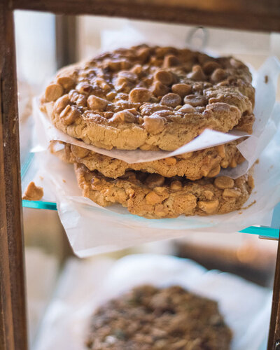 Large chewy cookies topped with peanut butter chips stacked in a bakery case at Gold Leaf Pastries in Kamloops BC.