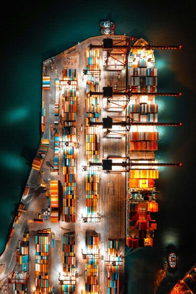 An aerial view of a brightly lit supply chain dock at night, featuring cranes and stacks of colorful shipping containers.