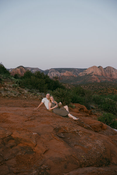a man and a woman snuggle together on red rocks against a backdrop of green brush landscaping and red rocks in the distance at sunset