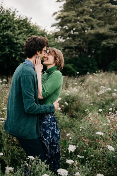 A woman holds her fiance close and leans in to kiss him as they stand in a wild flower patch in an Aberdeen park during their engagement session.