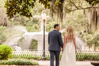 Eloping couple walking toward a fountain beneath oak trees with Spanish moss