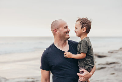 A father and son look at each other lovingly while they laugh together on the beach in San Diego