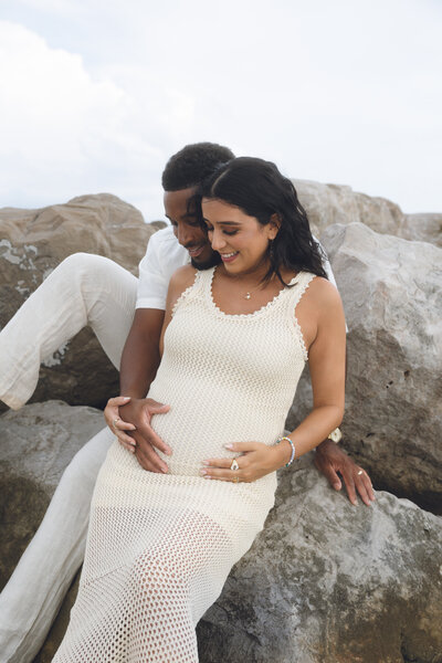 Expecting couple sitting on beach rocks, holding baby bump during maternity portrait session in New Jersey