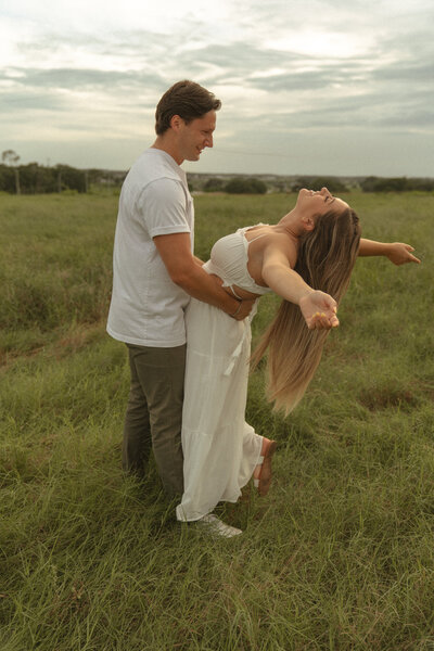 Man holding woman in a playful embrace as she leans back joyfully in an open grassy field at sunset.