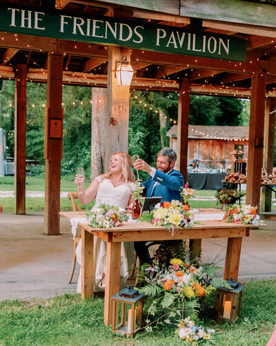 A couple toasts while sitting at a table, a sign above them saying "The Friends Pavilion."